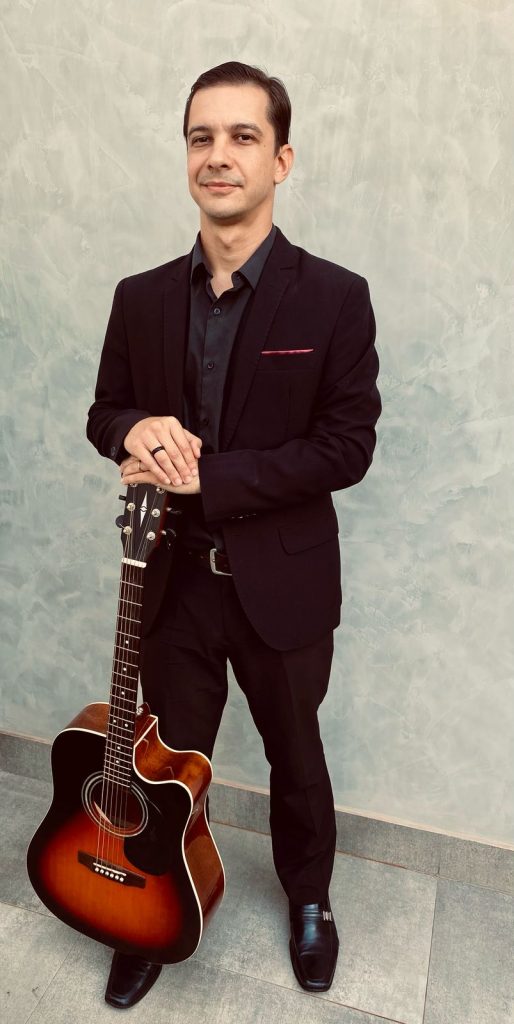 Músico Vini Roque de terno preto e segurando violão folk cort